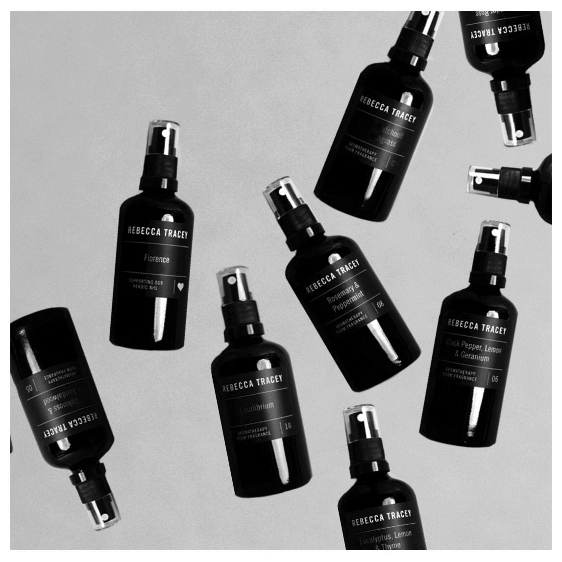 Multiple black spray bottles of Rebecca Tracey skincare products arranged diagonally on a light gray background, shown in a clean flat-lay composition.