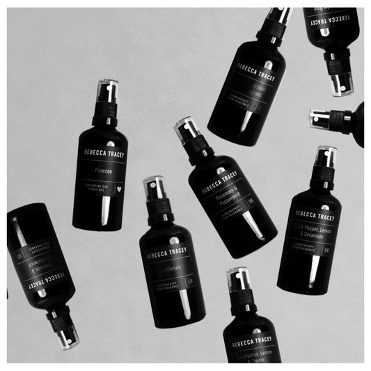 Multiple black spray bottles of Rebecca Tracey skincare products arranged diagonally on a light gray background, shown in a clean flat-lay composition.