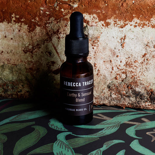 A small amber glass dropper bottle labeled Rebecca Trager Earthy & Sensual Blend Handmade Beard Oil, set against a rustic textured wall and a dark patterned surface with green tropical leaf motifs.