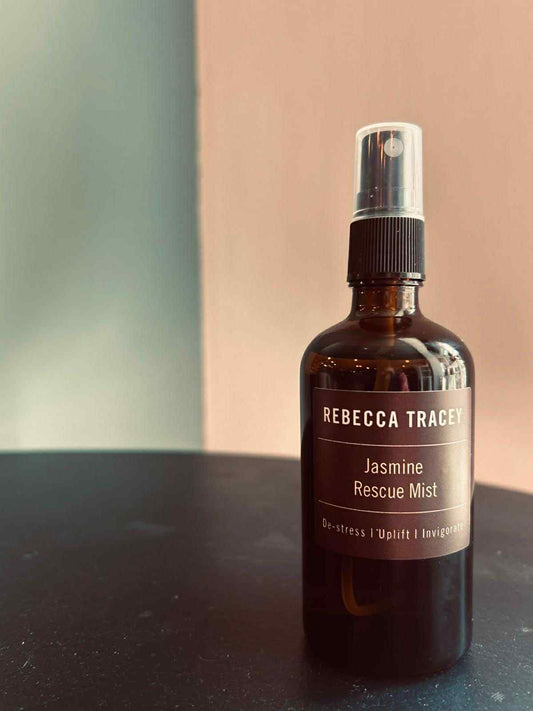 Brown spray bottle labeled 'Rebecca Tracey Jasmine Rescue Mist' on a dark surface with a blurred background.