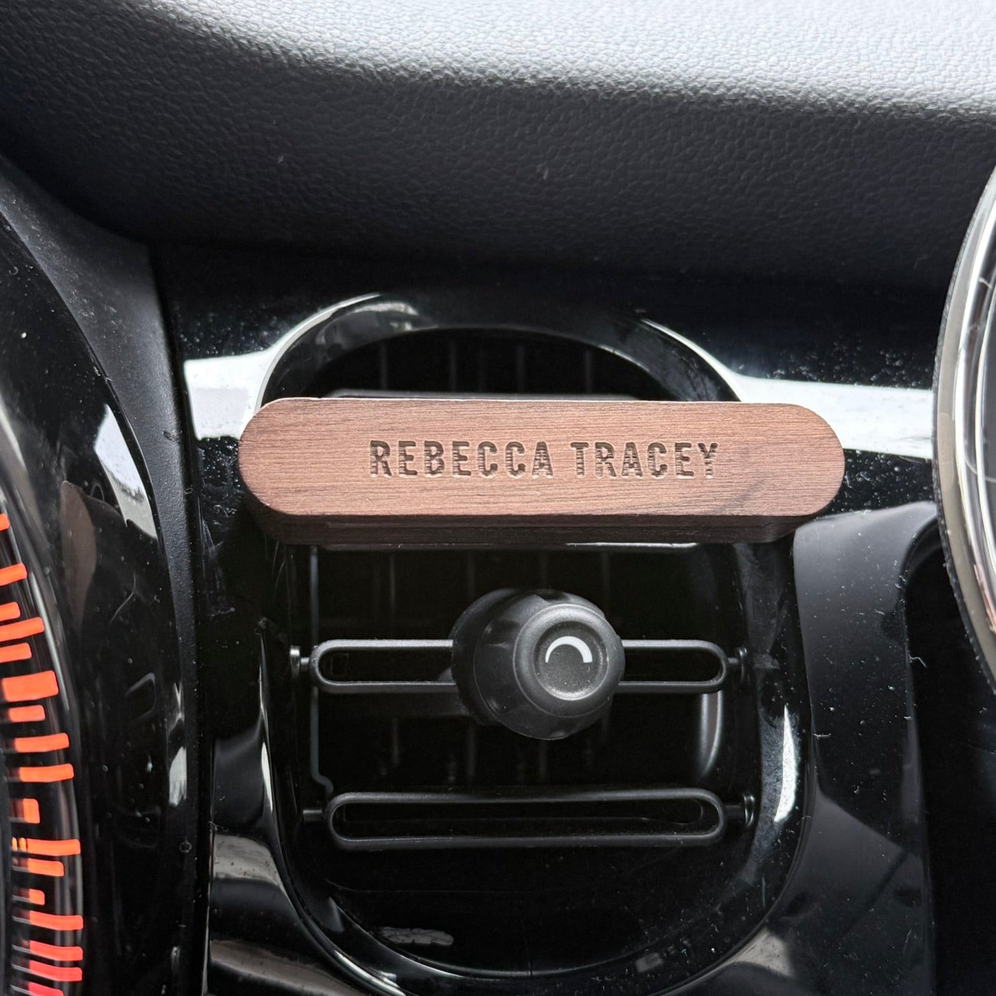 A personalized wooden car air freshener clip with the name 'REBECCA TRACEY' engraved, shown attached to a black car air vent.