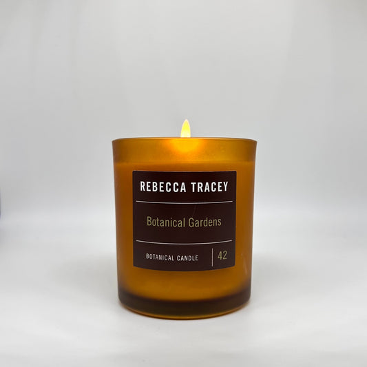 A lit amber glass candle with a dark brown label reading 'REBECCA TRACEY Botanical Gardens' on a light gray background.