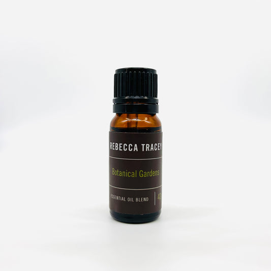 A small amber glass bottle of Rebecca Tracey Botanical Gardens essential oil blend with a black cap and dark brown label, centered on a white background.