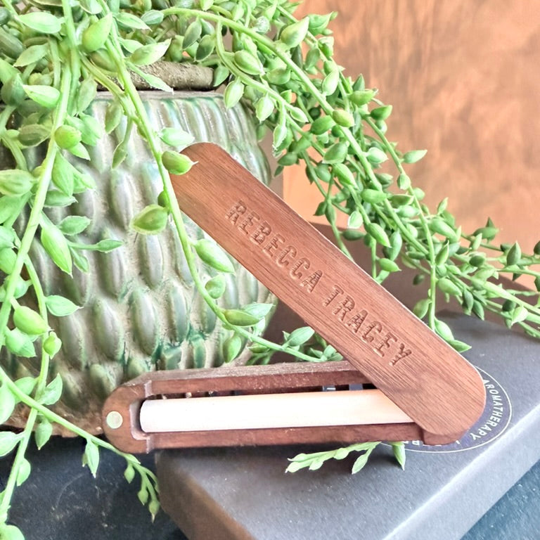 A wooden folding pocket knife with 'REBECCA TRACEY' engraved on the blade, resting on a dark gray book in front of a green trailing succulent plant.
