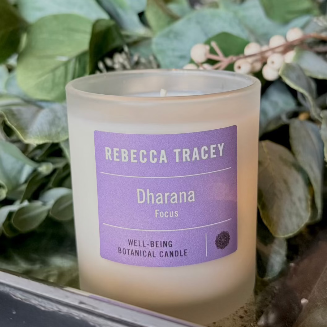 A Rebecca Tracey 'Dharana' botanical candle in a frosted glass jar, set against a background of green foliage and dried flowers.