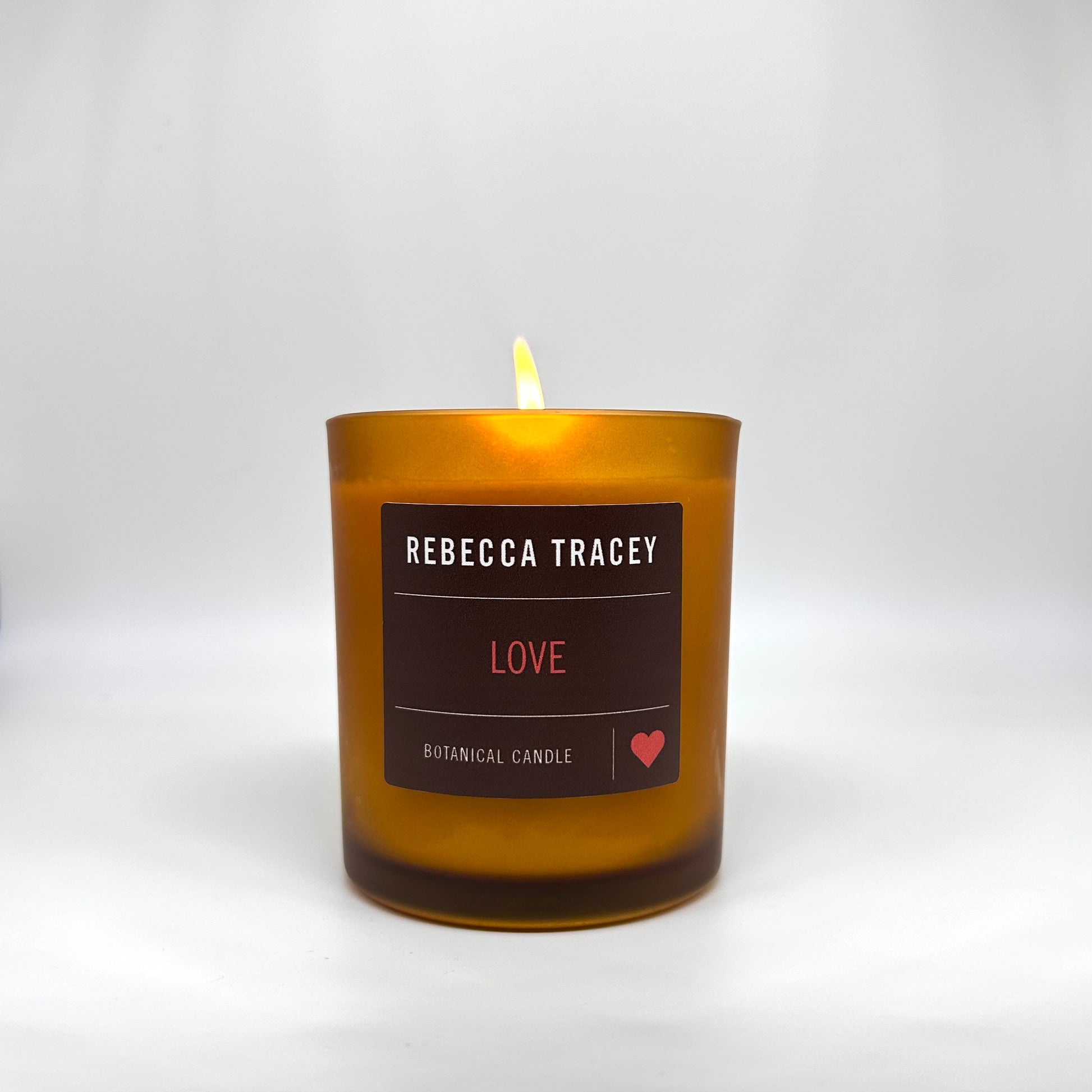 A lit amber glass candle with a dark brown label reading 'REBECCA TRACEY', 'LOVE', and 'BOTANICAL CANDLE' on a light gray background.