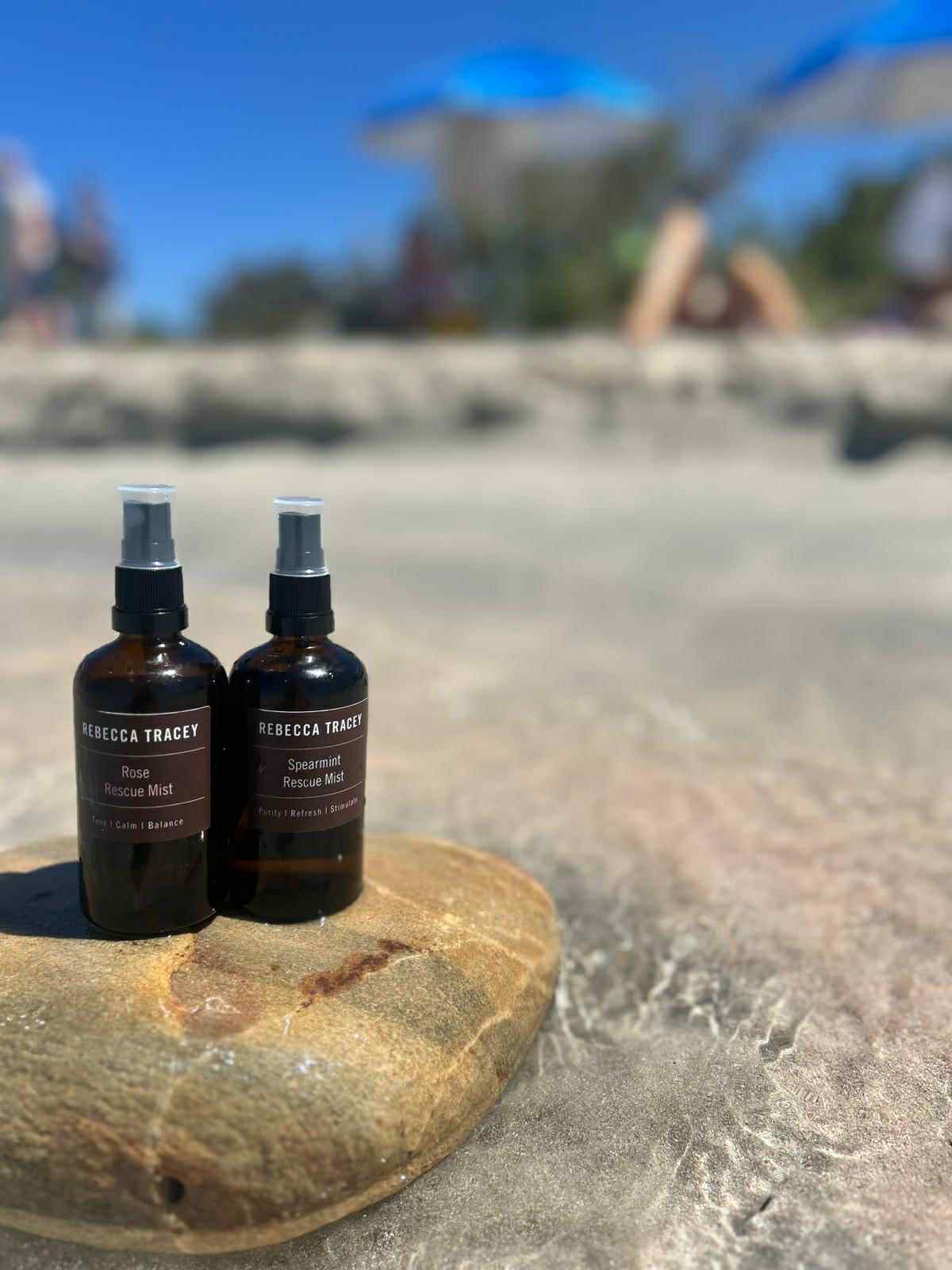 Two bottles of Rebecca Tracey products on a rock by a pool with people in the background