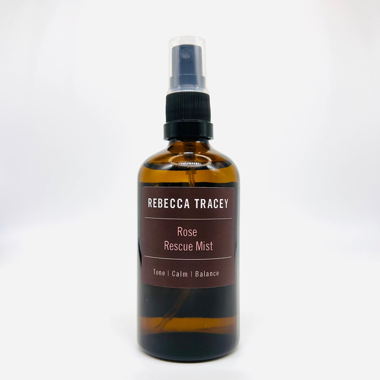 A bottle of Rebecca Tracey Rose Rescue Mist spray, featuring an amber glass bottle with a black spray nozzle and a dark brown label, set against a plain white background.