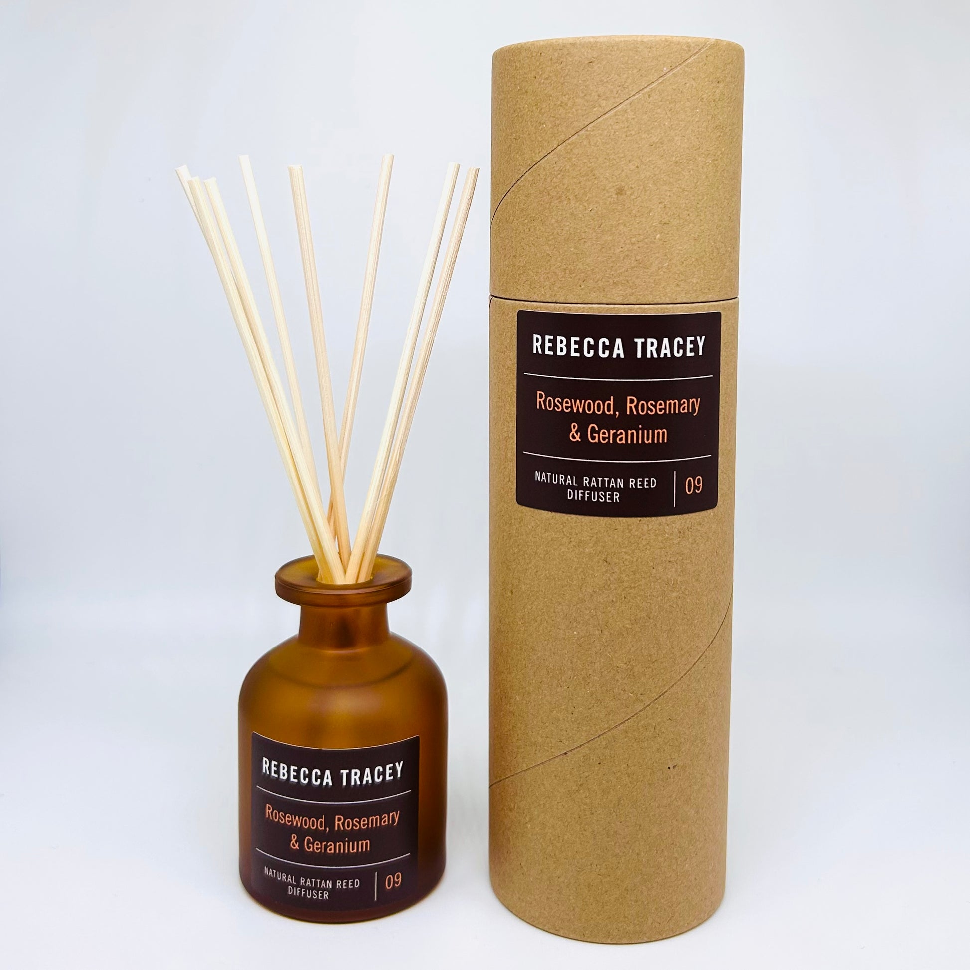 Amber glass reed diffuser bottle with natural reed sticks next to its brown kraft paper packaging cylinder, both labeled Rebecca Tracey Rosewood, Rosemary & Geranium.