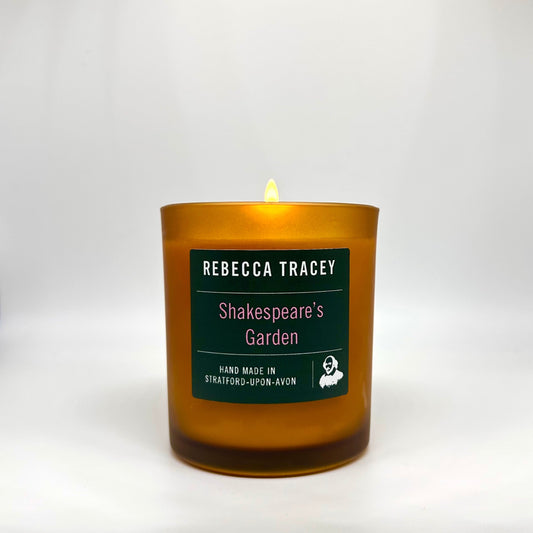 A lit amber glass candle with a dark green label reading 'REBECCA TRACEY Shakespeare's Garden' on a light gray background.