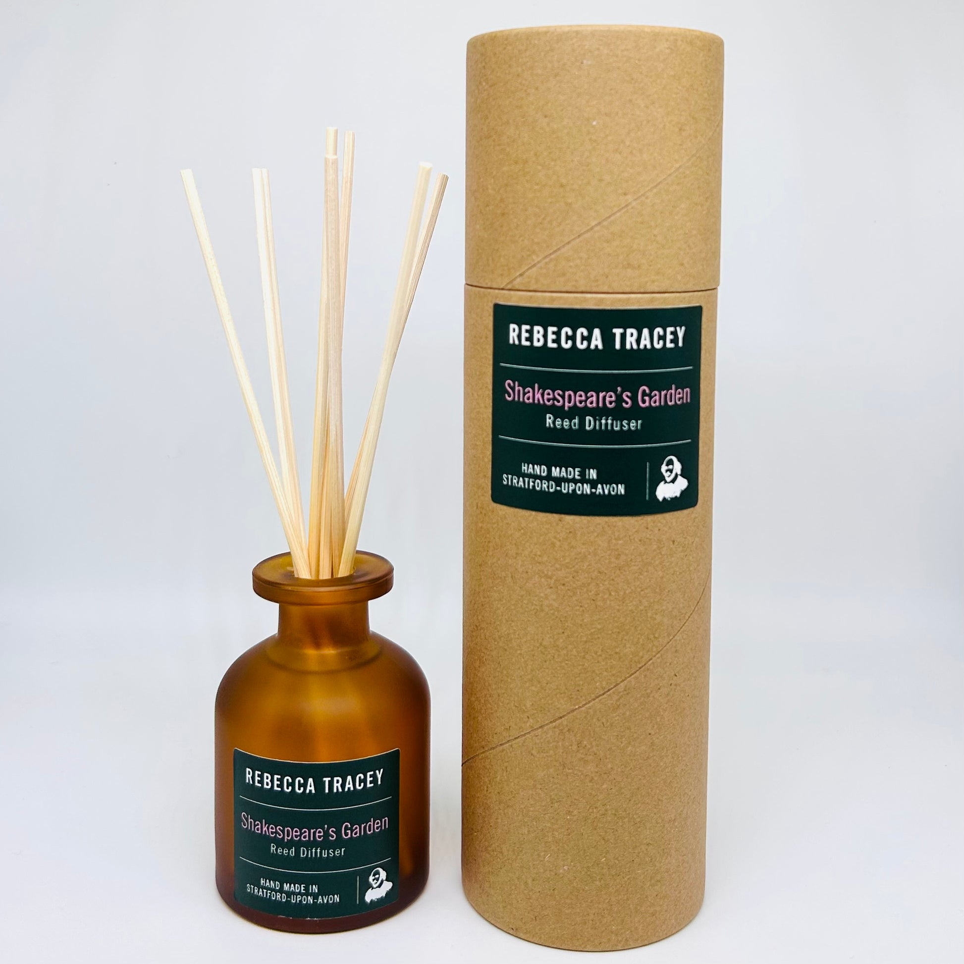 Rebecca Tracey Shakespeare's Garden Reed Diffuser set, featuring an amber glass bottle with natural reed sticks and its brown kraft paper cylindrical packaging box on a light gray background.