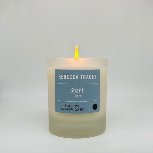 A lit Rebecca Tracey Shanti Peace botanical candle in a frosted glass jar, set against a neutral light gray background.