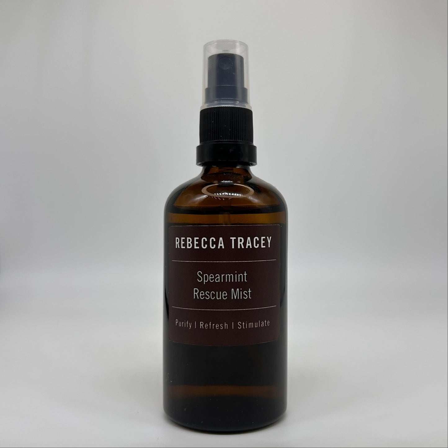 An amber glass spray bottle of Rebecca Tracey Spearmint Rescue Mist with a black spray cap and clear top, set against a neutral light gray background.