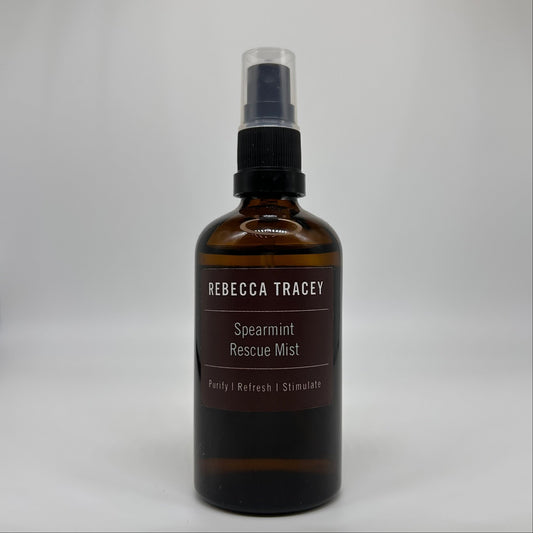 An amber glass spray bottle of Rebecca Tracey Spearmint Rescue Mist with a black spray cap and clear top, set against a neutral light gray background.