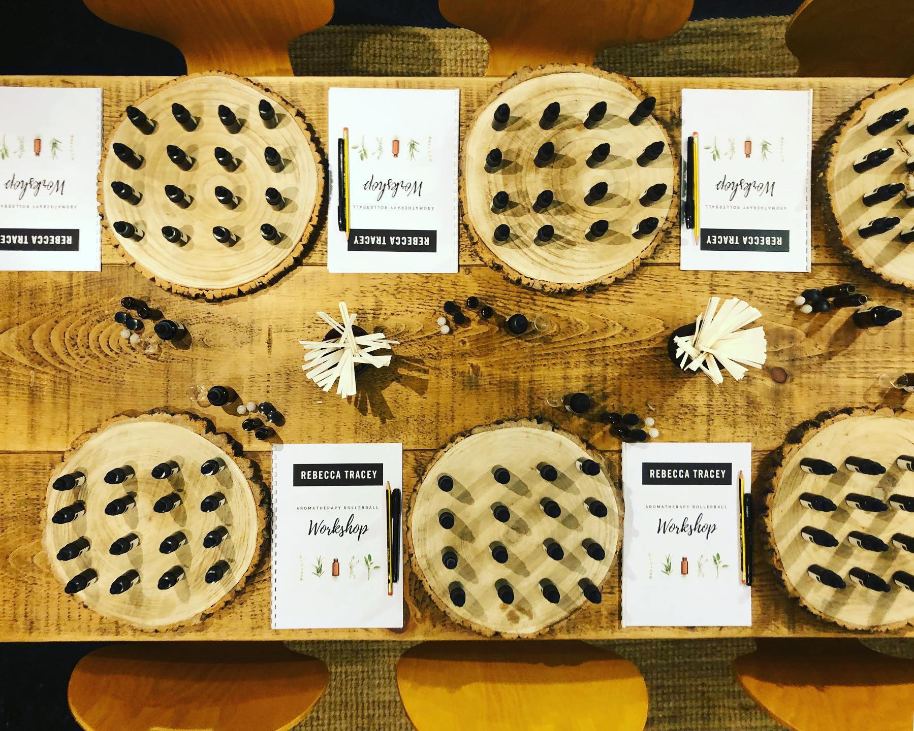 An overhead view of a wooden table displaying multiple round natural wood slices, each decorated with small black and white objects arranged in rows, alongside printed cards with names and workshop text.
