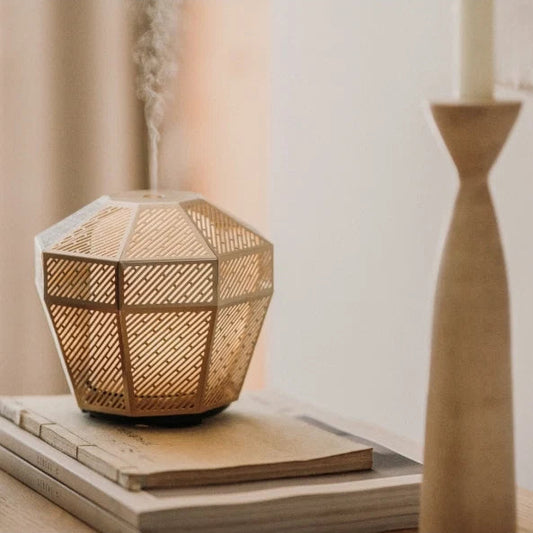 A faceted geometric diffuser with a woven natural rattan or wicker pattern, emitting a fine mist. It sits on a stack of books next to a wooden candlestick holder in a softly lit, warm-toned room.